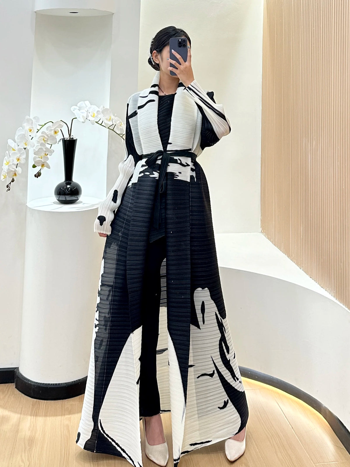 Une femme modélise un trench-coat à manches longues à plis noir et blanc dans un intérieur lumineux.