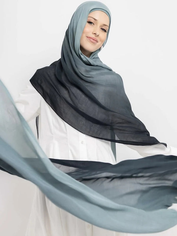 A woman wearing a gray hijab and a black scarf with a sheer blue fabric flowing around her.