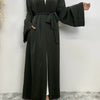 Eine Frau in einem langen, dunkelgrünen, fließenden Abaya-Kaftan mit offenen Front und gebundener Taille vor einer weißen Wan