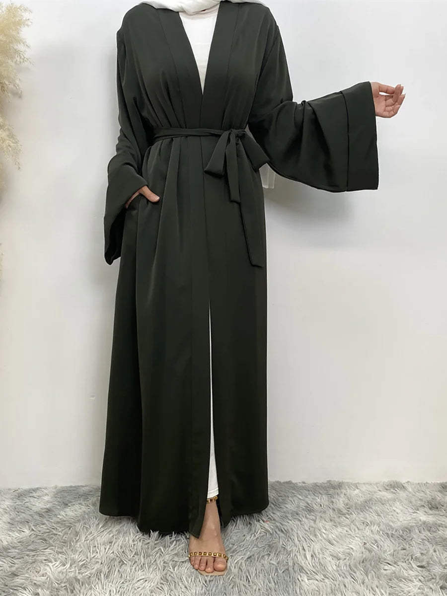 Eine Frau in einem langen, dunkelgrünen, fließenden Abaya-Kaftan mit offenen Front und gebundener Taille vor einer weißen Wan