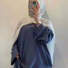 Eine Frau trägt eine blaue, fließende, bodenlange Abaya mit weiten Ärmeln und einen weißen Hijab und hält ein Smartphone.