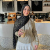 Eine Frau mit schwarzem Hirschmuster-Hijabschal und beigem Oberteil hält ein Getränk in einem Café.