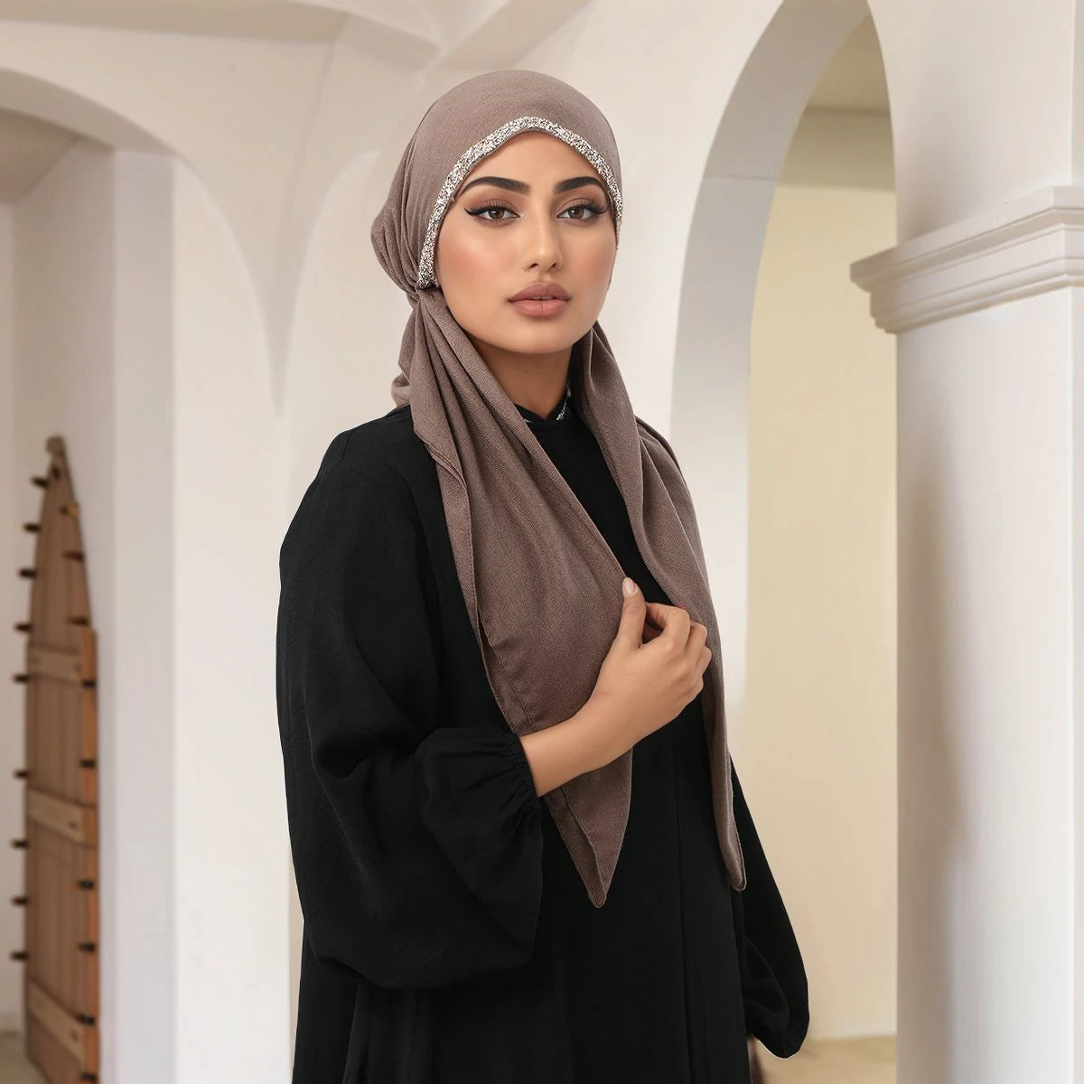 Une femme vêtue de noir et d'un hijab taupe orné de diamants, debout dans un couloir architectural.