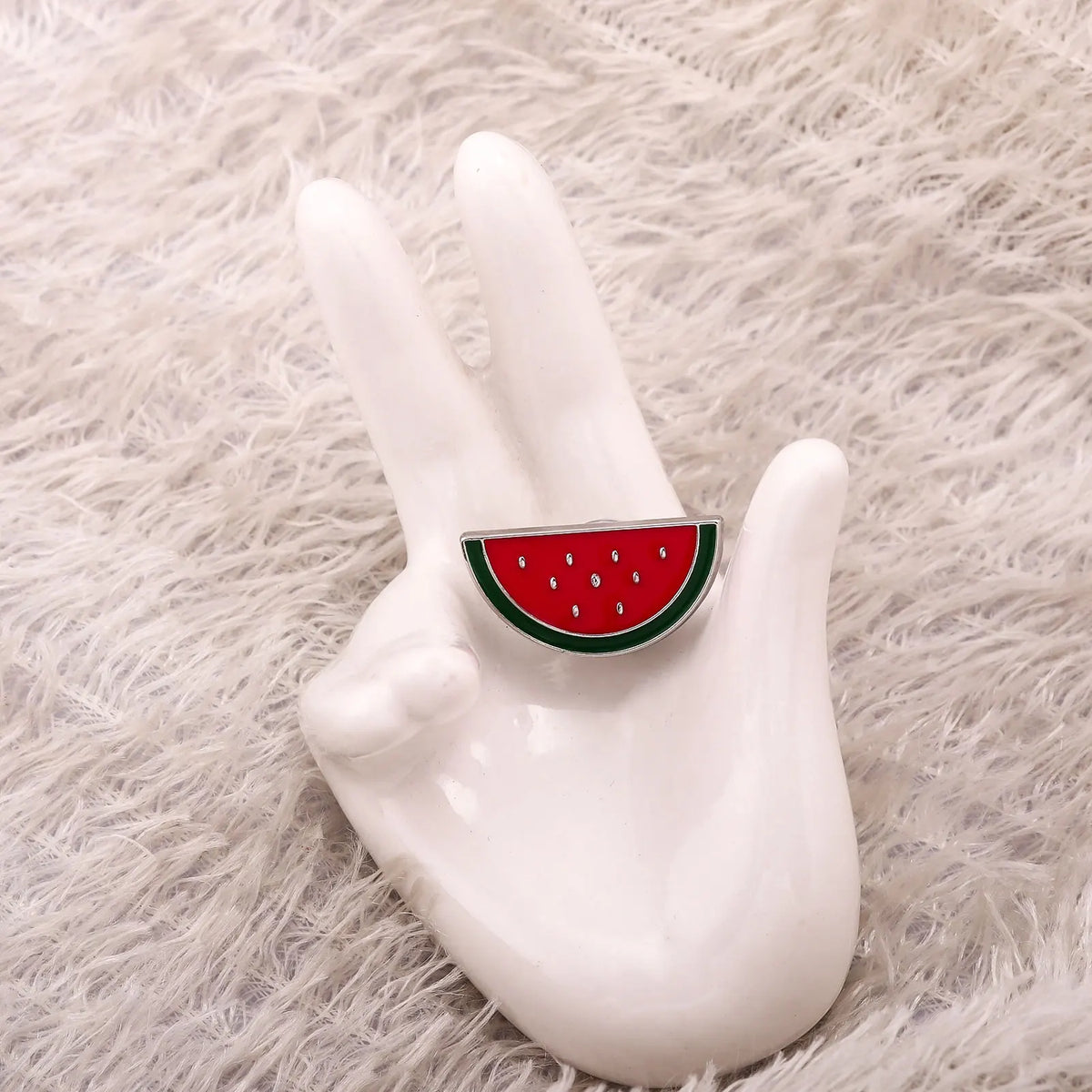 Weißes Emaille-Reversspange in Form einer Wassermelonen-Scheibe auf weißem flauschigem Fell.