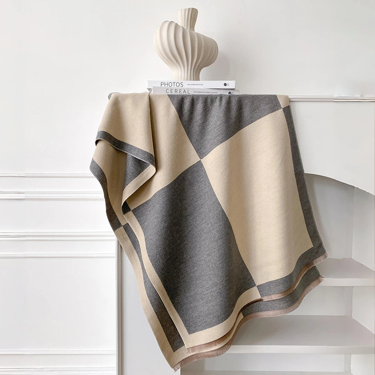 Pañuelo pashmina geométrico beige y gris sobre una estantería blanca con un jarrón de cerámica.