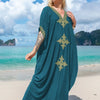 Eine Frau in einem teal-gold-bestickten V-Ausschnitt-Maxikaftan steht auf einem Sandstrand mit Ozeangewällen im Hintergrund.