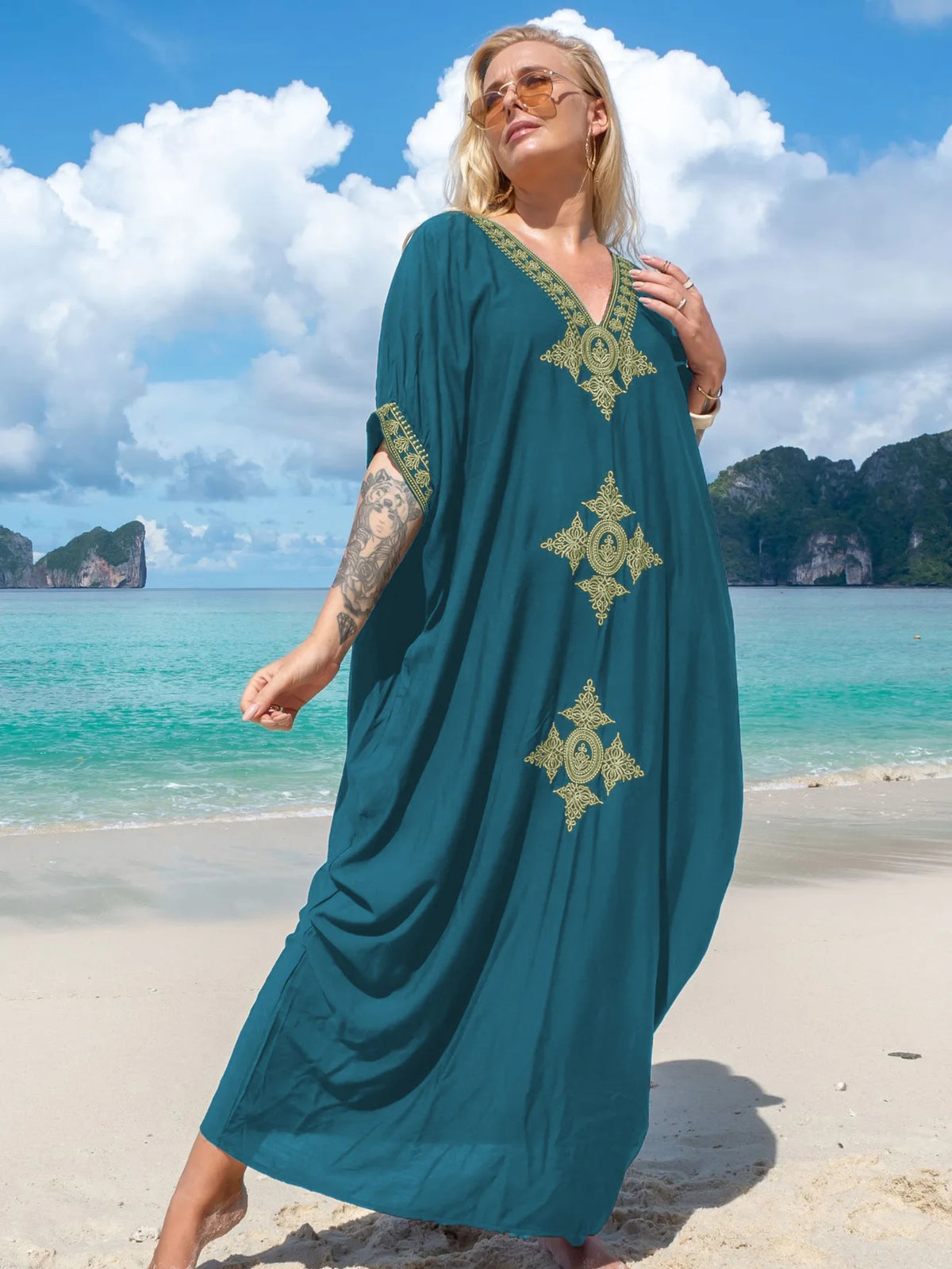 Eine Frau in einem teal-gold-bestickten V-Ausschnitt-Maxikaftan steht auf einem Sandstrand mit Ozeangewällen im Hintergrund.