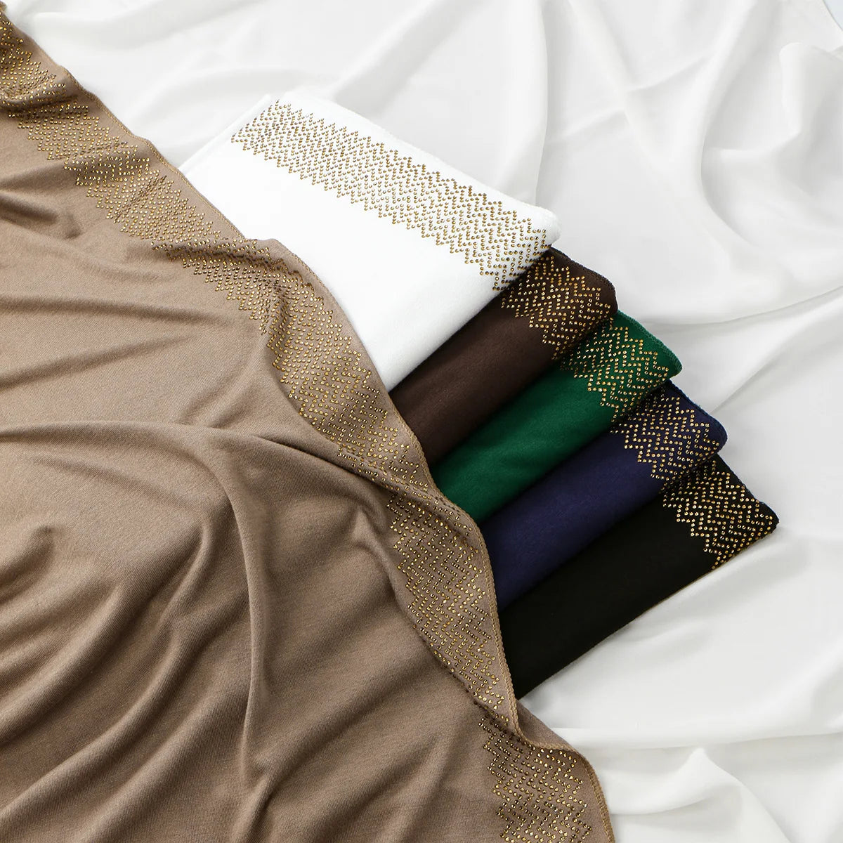 Ein Stapel funkelnder Strass-Hijabs in verschiedenen Erdtönen und dunklen Farben, drapiert vor weißem Hintergrund