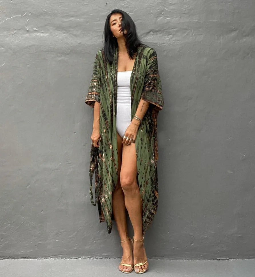 Eine Frau trägt ein langes, olivgrünes Blumenkimono-Überkleid über einem weißen Kleid vor einer grauen Wand.