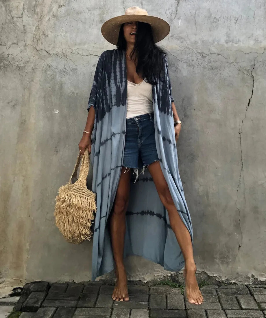 Eine Frau in blau-weiß gemustertem Kimono-Überwurf über einem weißen Top und Jeansshorts vor einer Betonwand.