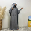 Une personne portant une abaya grise avec capuche et manches batwing se tient contre un mur blanc.