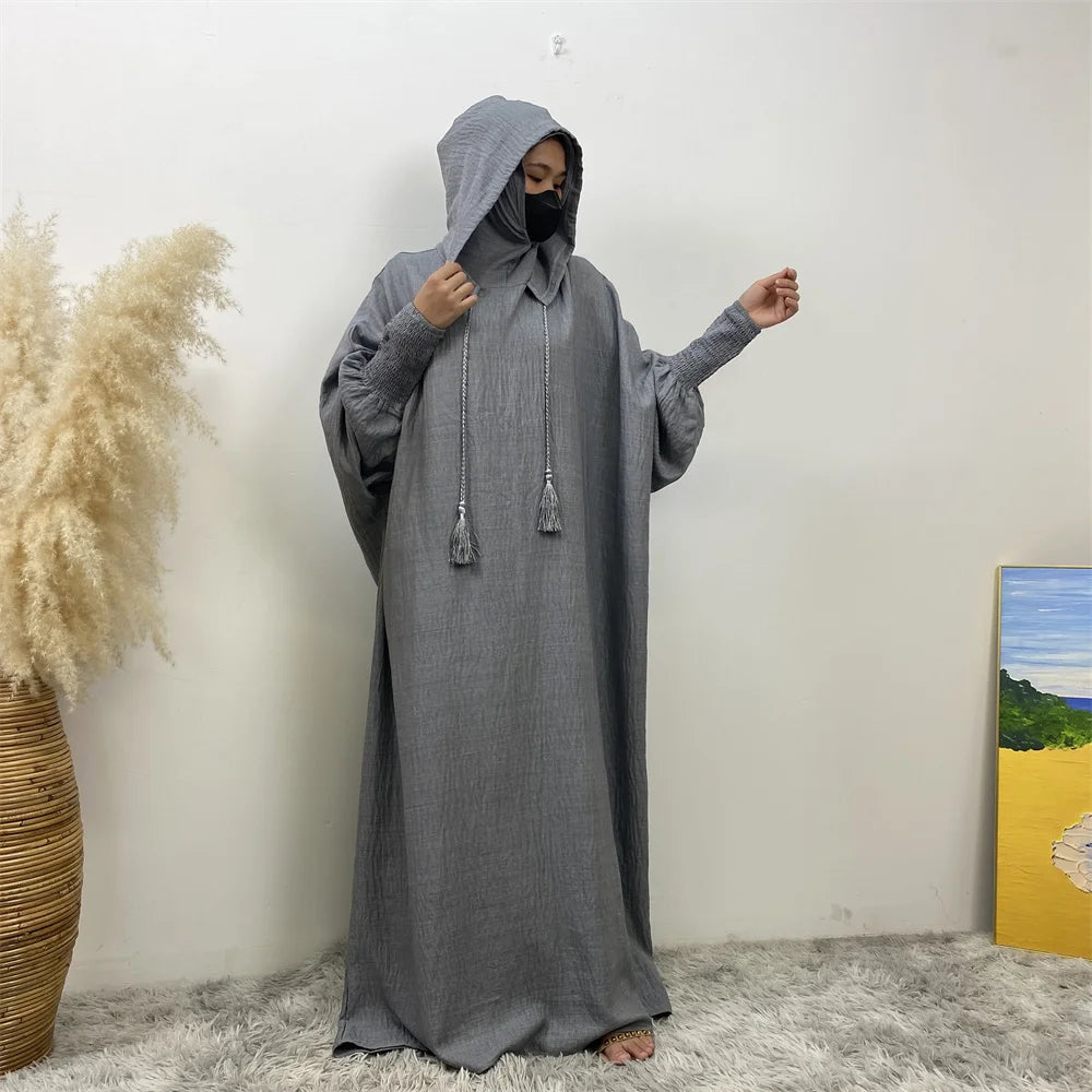 Une personne portant une abaya grise avec capuche et manches batwing se tient contre un mur blanc.