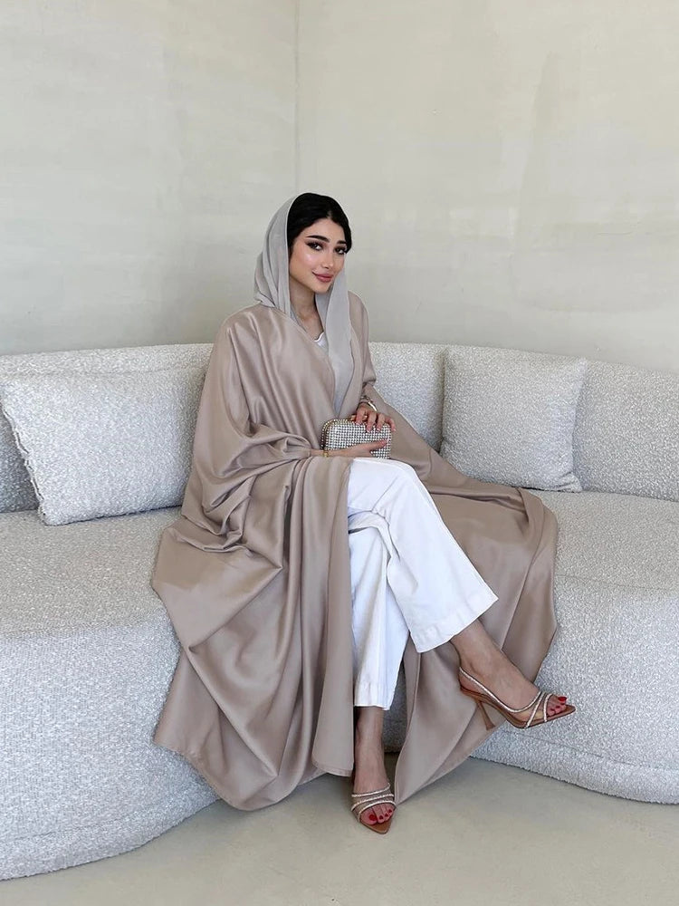Una mujer vistiendo una abaya de satén beige con mangas de murciélago y pantalones blancos, sentada en un sofá.