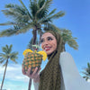 Eine Frau mit olivgrünem Polka-Dot-Hijab hält eine Ananas vor einem leuchtend blauen Himmel mit Palmen.