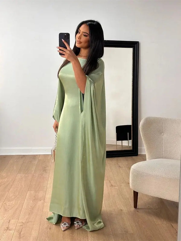 A woman models a sage green satin batwing abaya in a full-length dress in a bright room.