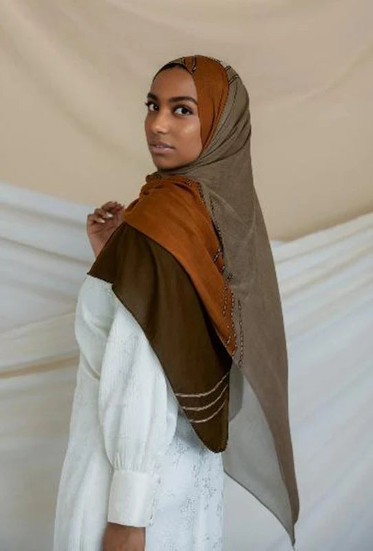 Una mujer vistiendo un pañuelo de hijab marrón y beige sobre una blusa blanca.