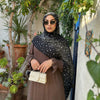Eine Frau mit dunklem gemustertem Hijab-Schal und Sonnenbrille steht draußen vor einer strukturierten Wand mit Grünpflanzen.