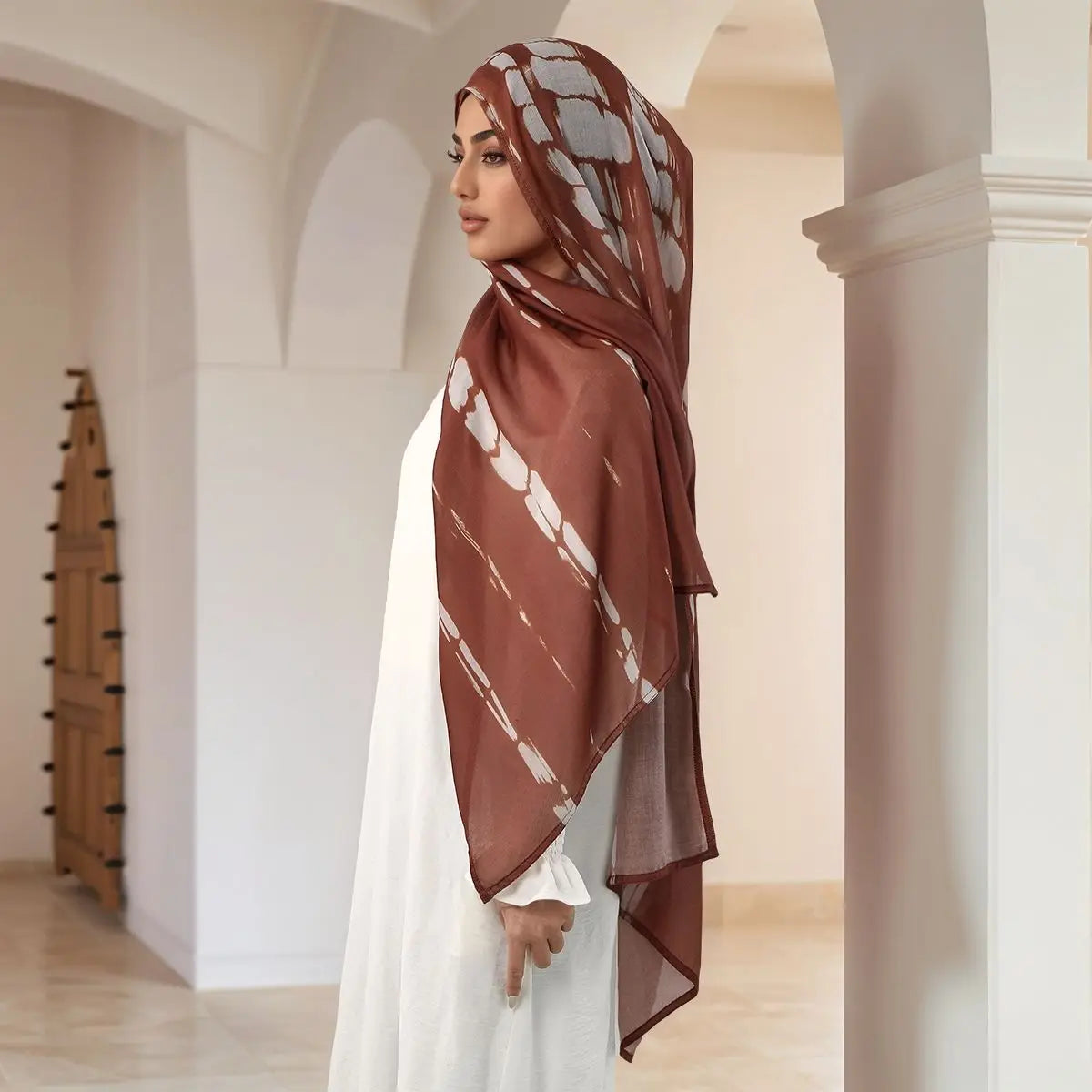 A woman wearing an elegant printed linen hijab scarf in brown and white tones stands in a bright interior.