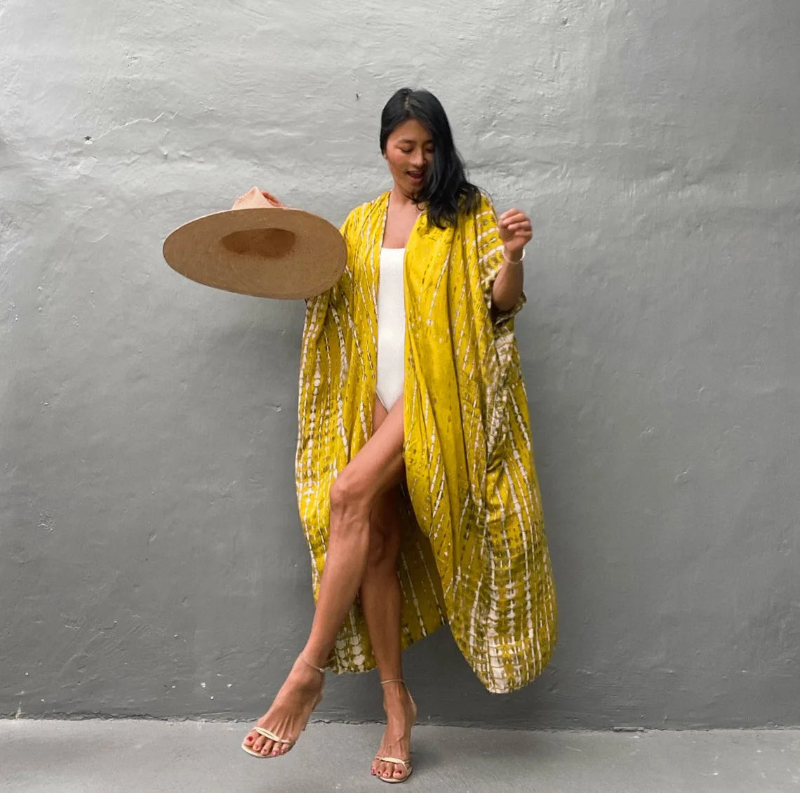 Eine Frau in einem gelb gemusterten Kimono-Überkleid und einem breiten Strohhut vor einer grauen Wand.