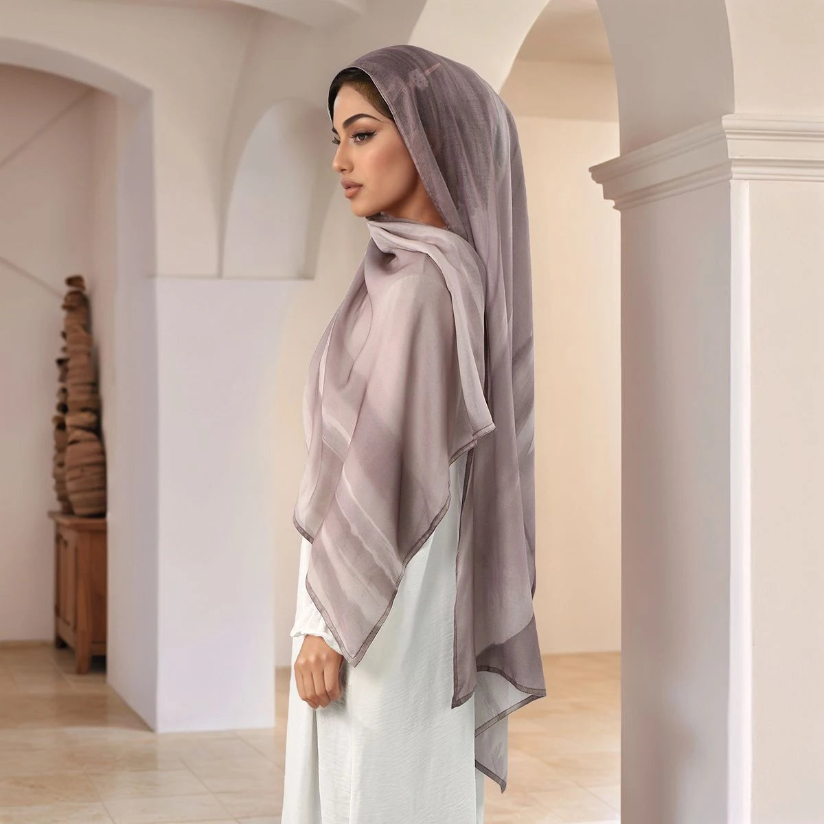 Eine Frau in einem hellgrauen bedruckten Baumwoll-Rayon-Hijab und weißem Kleid in einem gewölbten Flur.