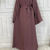 Elegane mauve maxi Abaya für Frauen hängt an einer weißen Paneelwand.
