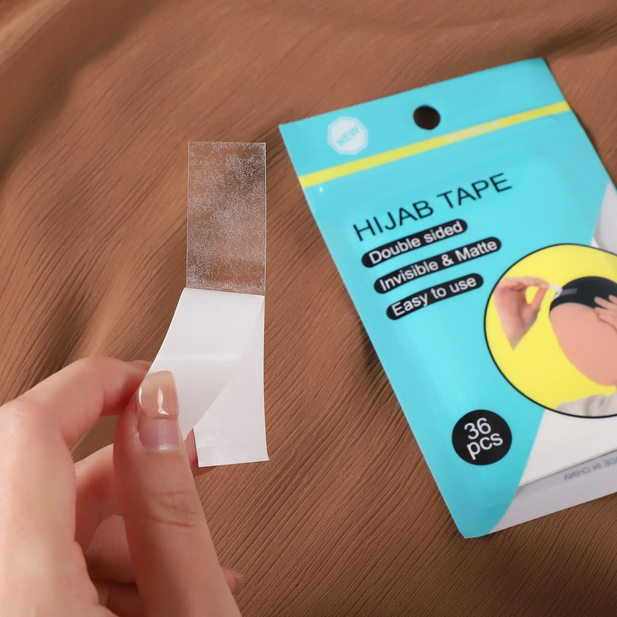 Una mano aplica cinta de doble cara transparente a un trozo de tela blanca, junto al empaque del producto.