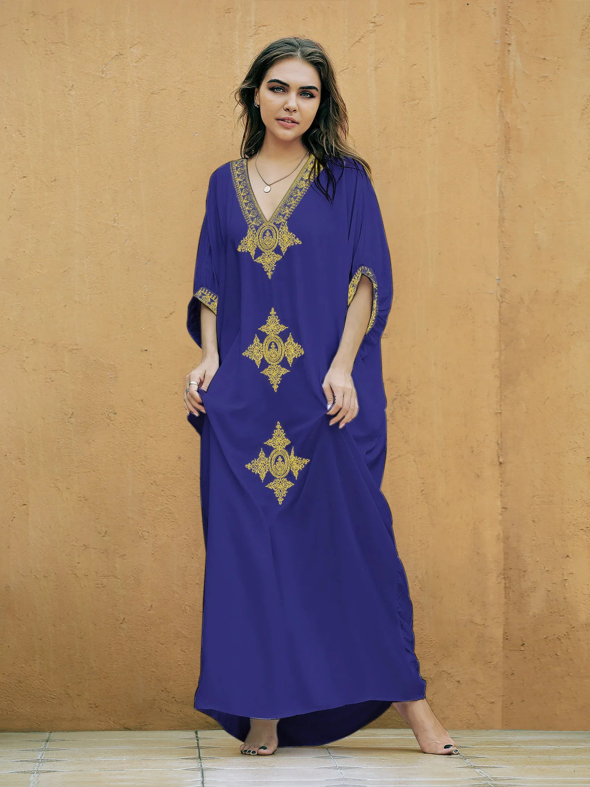 Eine Frau modelliert ein lila Kaftankleid mit goldener Stickerei vor einer orangefarbenen Wand.
