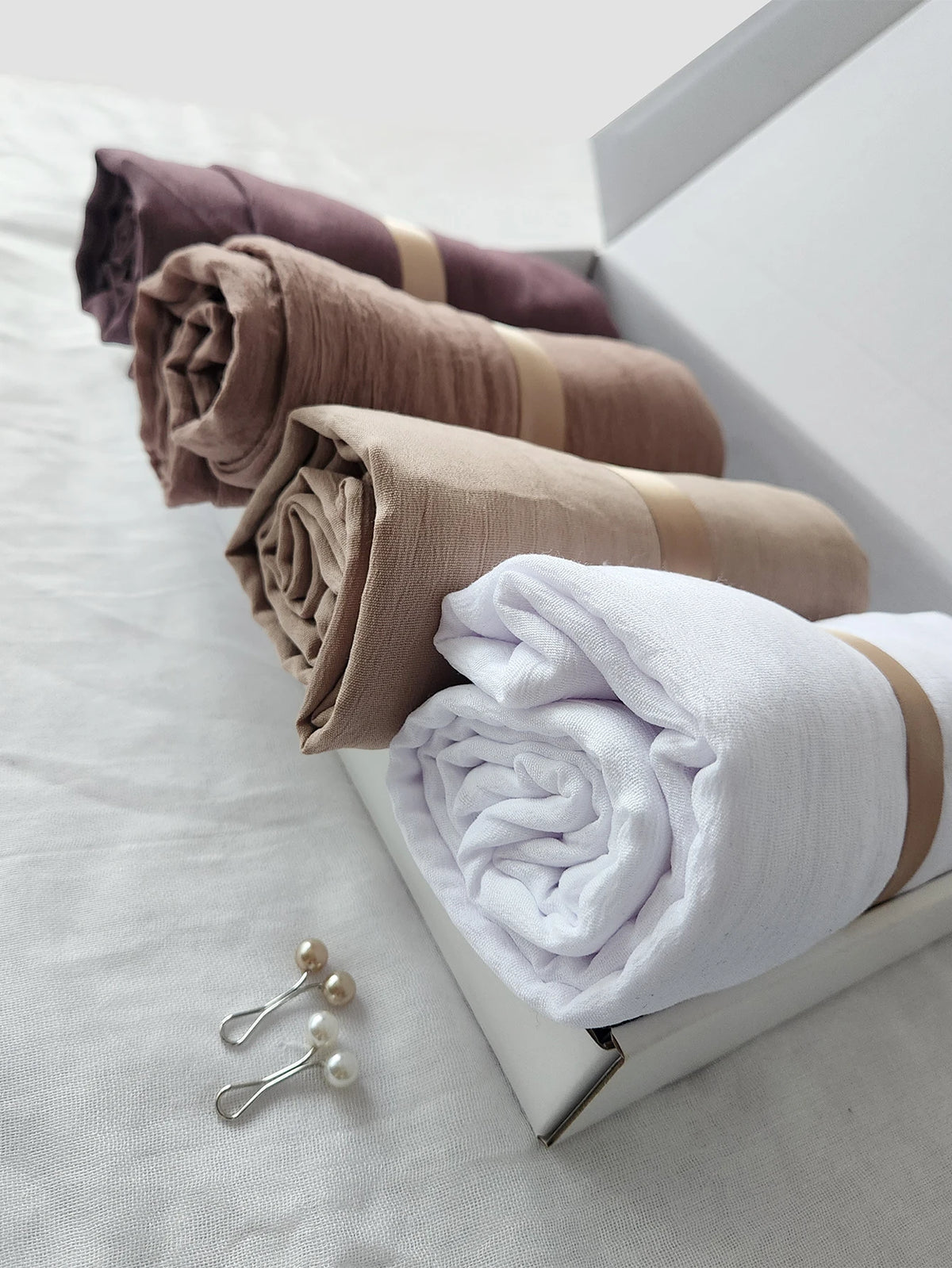 Ein Set aus vier zerknitterten Baumwoll-Hijab-Schals in Mauve-, Beige- und Weißtönen, gestapelt in einer Box.