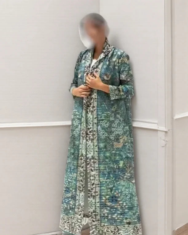 Una mujer vistiendo un cárdigan abaya plisado de color turquesa y blanco, de pie en un marco de puerta.