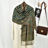 Chal de bordado paisley en verde azulado oscuro y dorado sobre una prenda blanca, junto a un bolso de cuero marrón.
