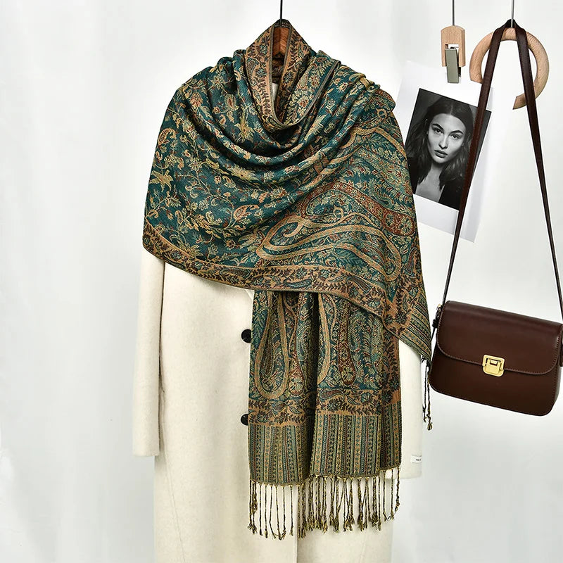 Chal de bordado paisley en verde azulado oscuro y dorado sobre una prenda blanca, junto a un bolso de cuero marrón.