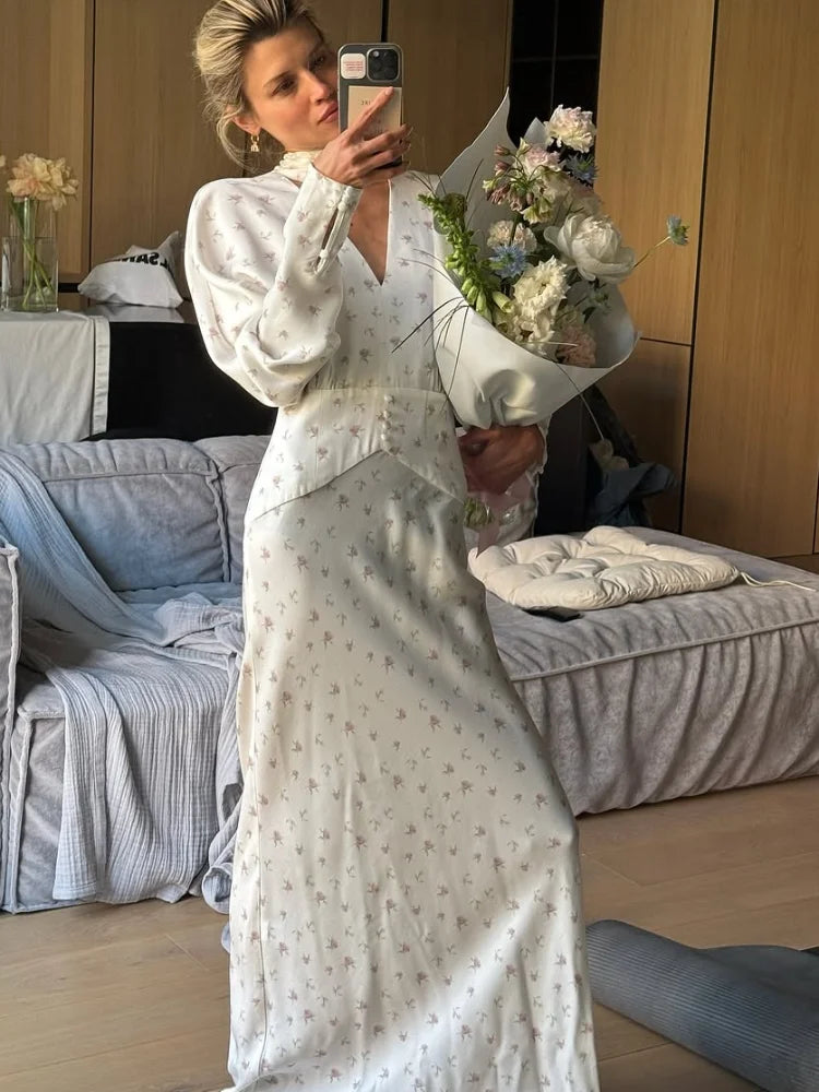 Una mujer viste un vestido largo de satén con estampado floral blanco y está en un dormitorio moderno.