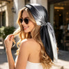 Une femme blonde portant un foulard dégradé noir et argent autour de ses cheveux, souriante à l'extérieur.