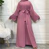 A woman wearing a long, flowing dusty rose abaya and kaftan set in a light-colored room.