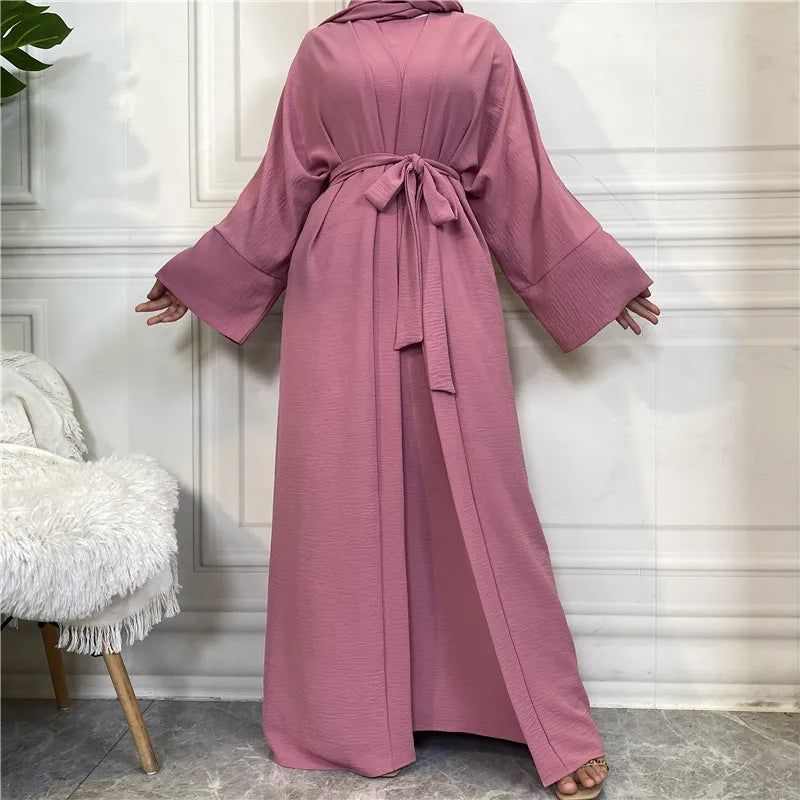 A woman wearing a long, flowing dusty rose abaya and kaftan set in a light-colored room.