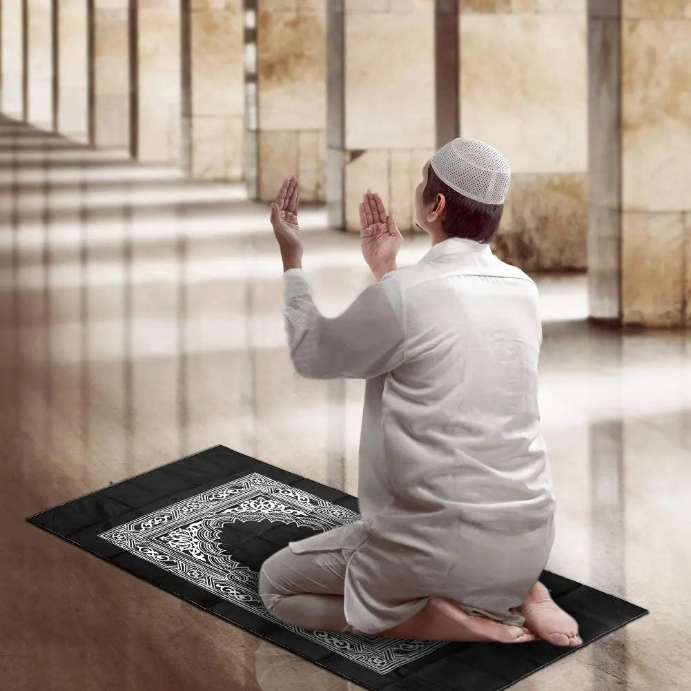 A person in a white shirt is sitting on a prayer rug with hands raised in a prayer gesture.