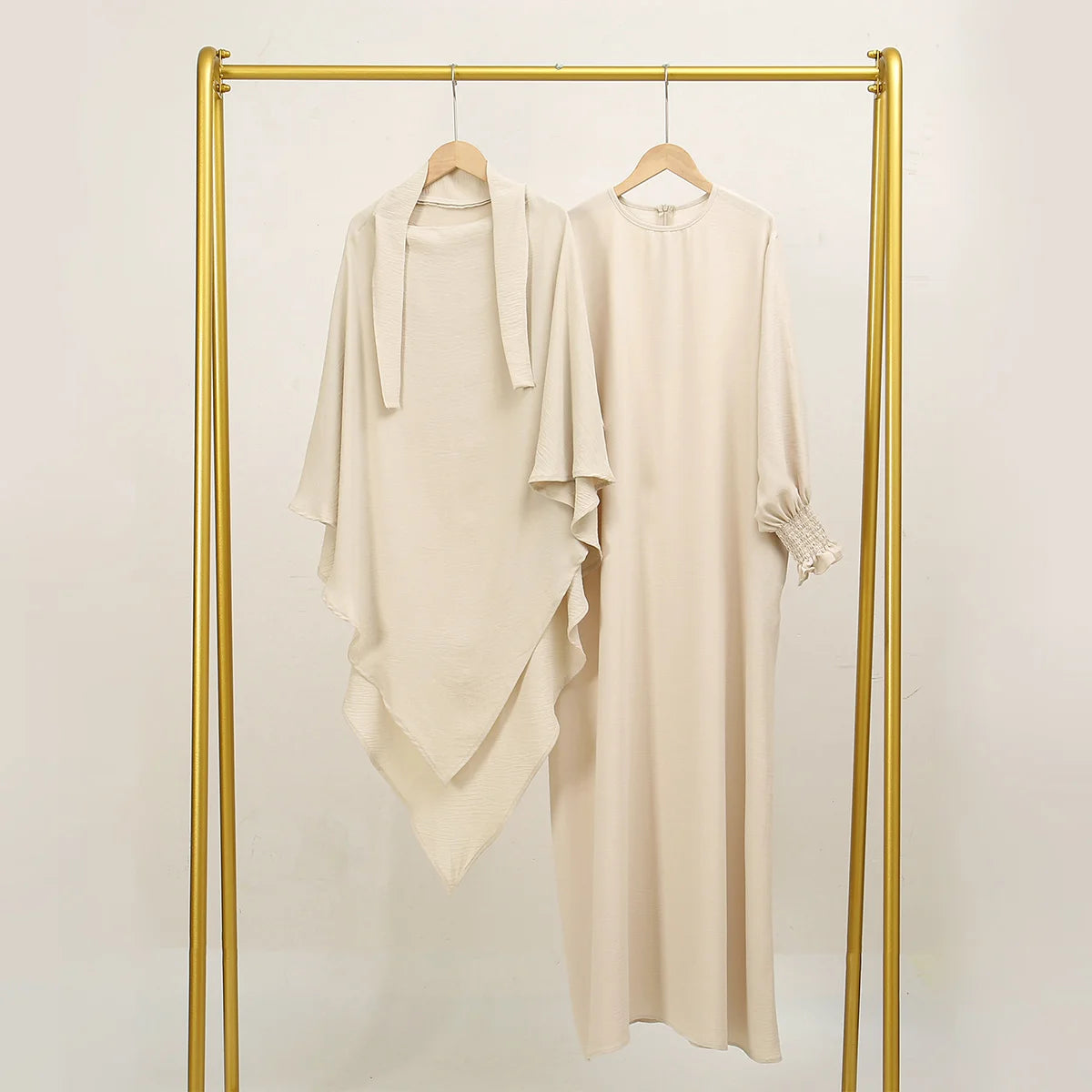 Beige elegante Abaya und Khimar-Set auf goldener Kleiderstange vor weißem Hintergrund.