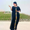 Eine Frau in einem schwarzen Kaftan mit farbenfroher Stickerei und Sonnenbrille steht vor einem grünen Hügel.
