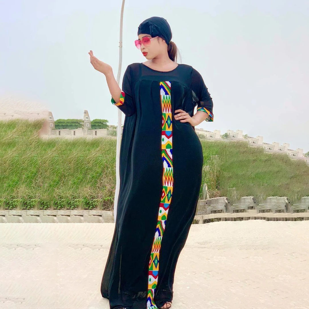 Eine Frau in einem schwarzen Kaftan mit farbenfroher Stickerei und Sonnenbrille steht vor einem grünen Hügel.