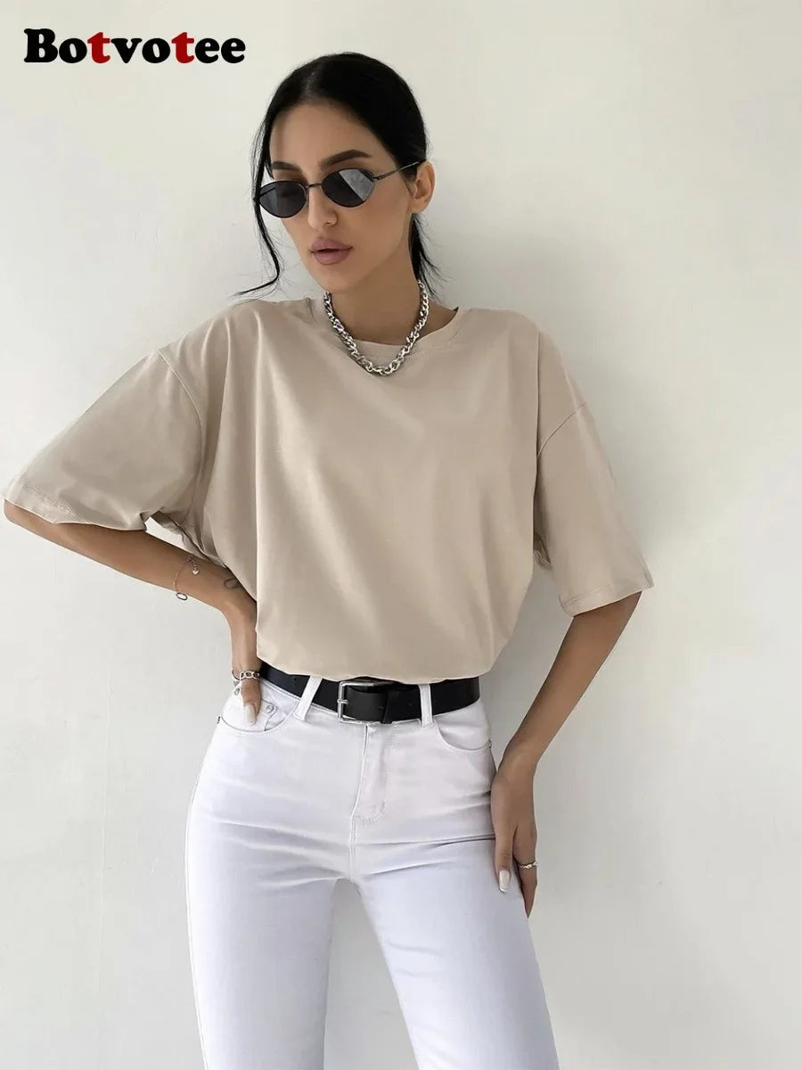 Una mujer modela una camiseta básica de algodón beige oversize, jeans blancos y un collar de plata.