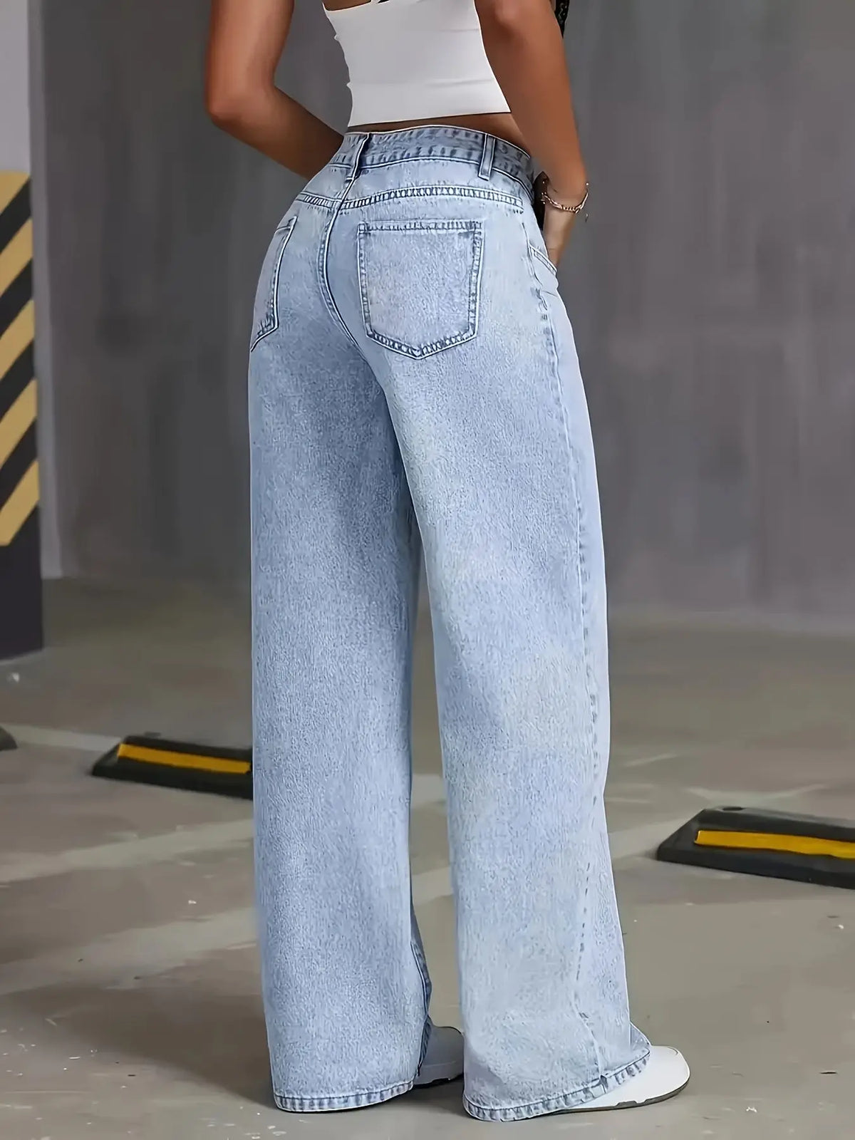 Una mujer vistiendo pantalones vaqueros anchos azul claro y una blusa blanca, de pie en interiores.