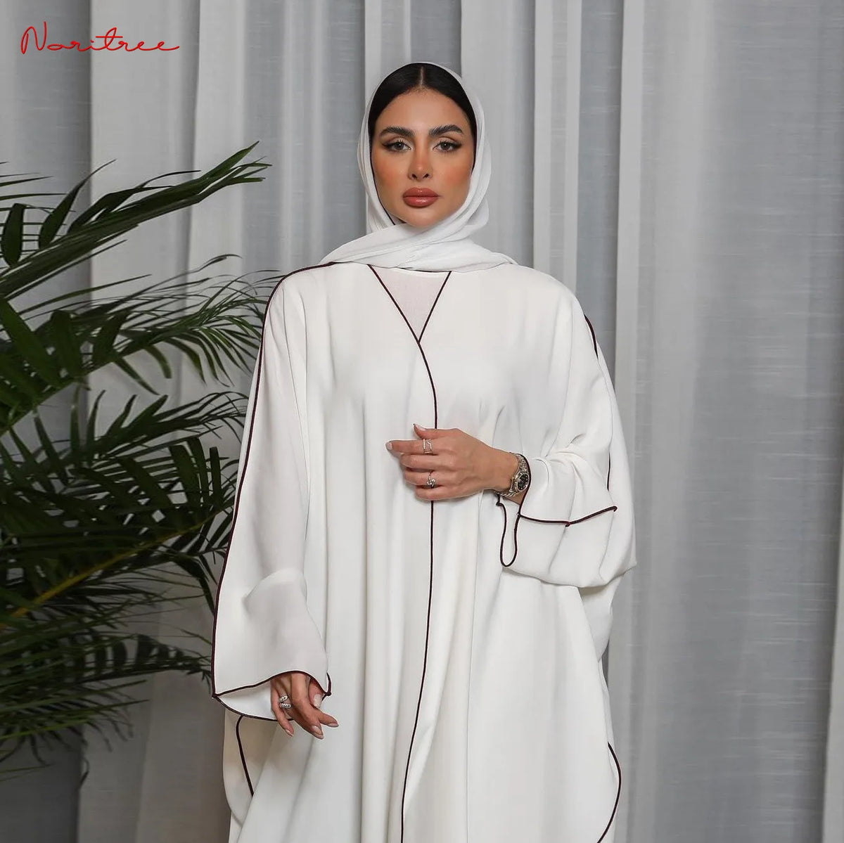 Una mujer vistiendo una abaya blanca oversize con ribetes marrones y un hiyab a juego, de pie en interiores.