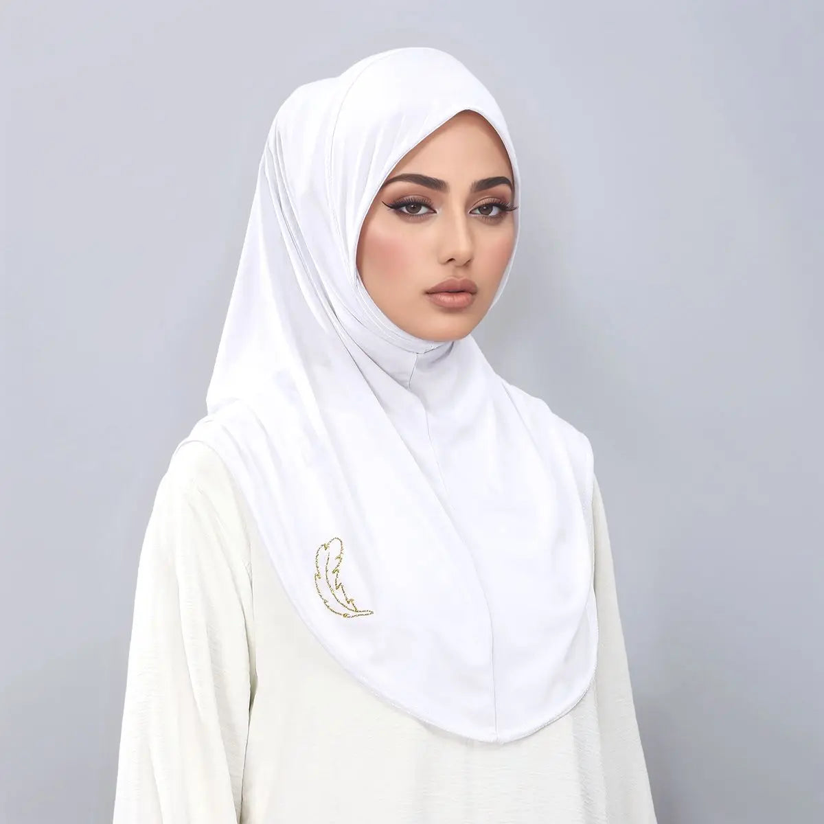 Une femme portant un hijab blanc avec broderies dorées sur un fond gris clair.