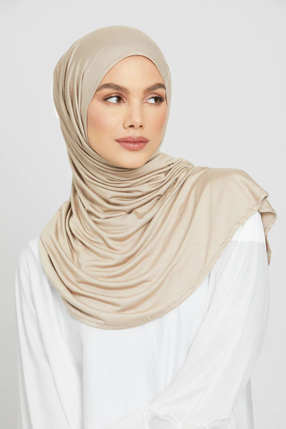 Eine Frau trägt einen beige Jersey-Hijab und ein weißes Oberteil.
