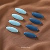 Eight oval blue brooch pins arranged on a brown fabric background.