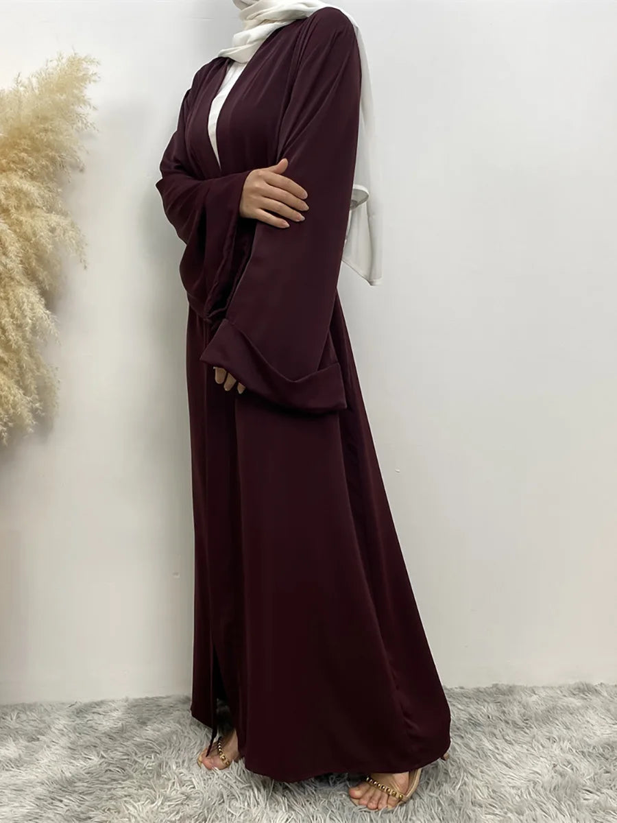 Eine Frau in einem langen, fließenden dunkelroten Abaya-Kaftan mit weißem Hijab vor hellem Hintergrund.