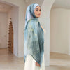 A woman wearing an elegant printed linen hijab scarf in blue and white tones, standing indoors.