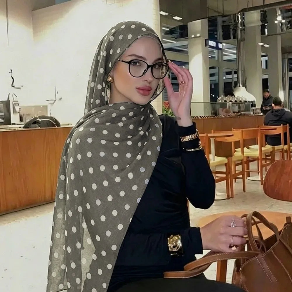 Eine Frau mit grauem Polka-Dot-Baumwollschal und Brille sitzt an einem Holztisch in einem modernen Café.