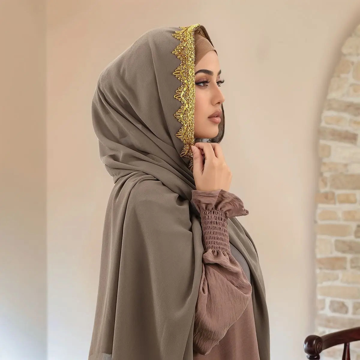 Eine Frau trägt einen beige-zarten Chiffon-Hijabschal mit goldenem Zierrand und ein braunes Oberteil.