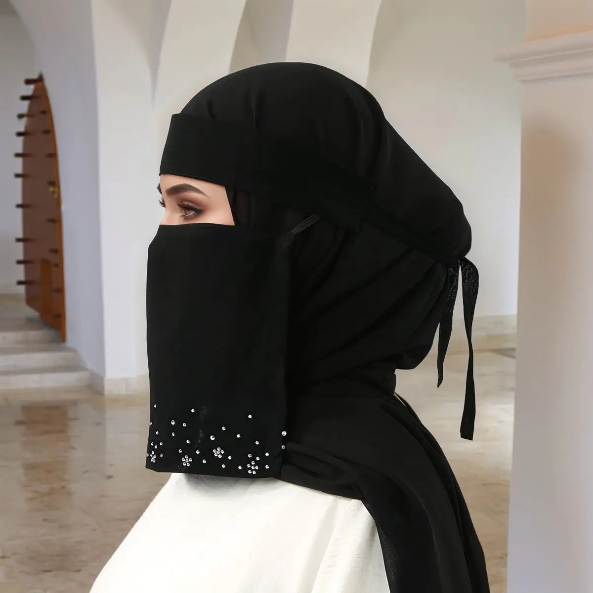 Eine Frau trägt einen eleganten schwarzen Hijab mit floralen Diamantdetails und einem Gesichtsveil und blickt zur Seite.
